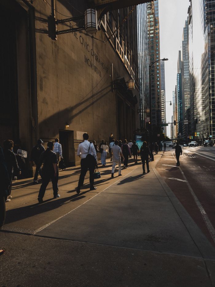Pedestrians walking on a sunlit street in a bustling downtown city, surrounded by skyscrapers.